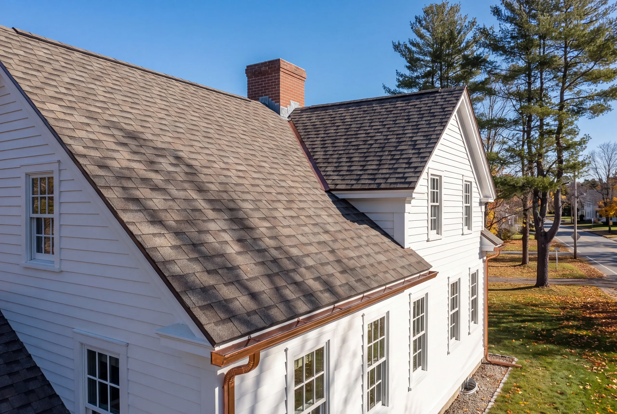 Residential roofing contractor Pittsfield Maine: white house with dark shingles, brick chimney and copper gutters in an autumn yard