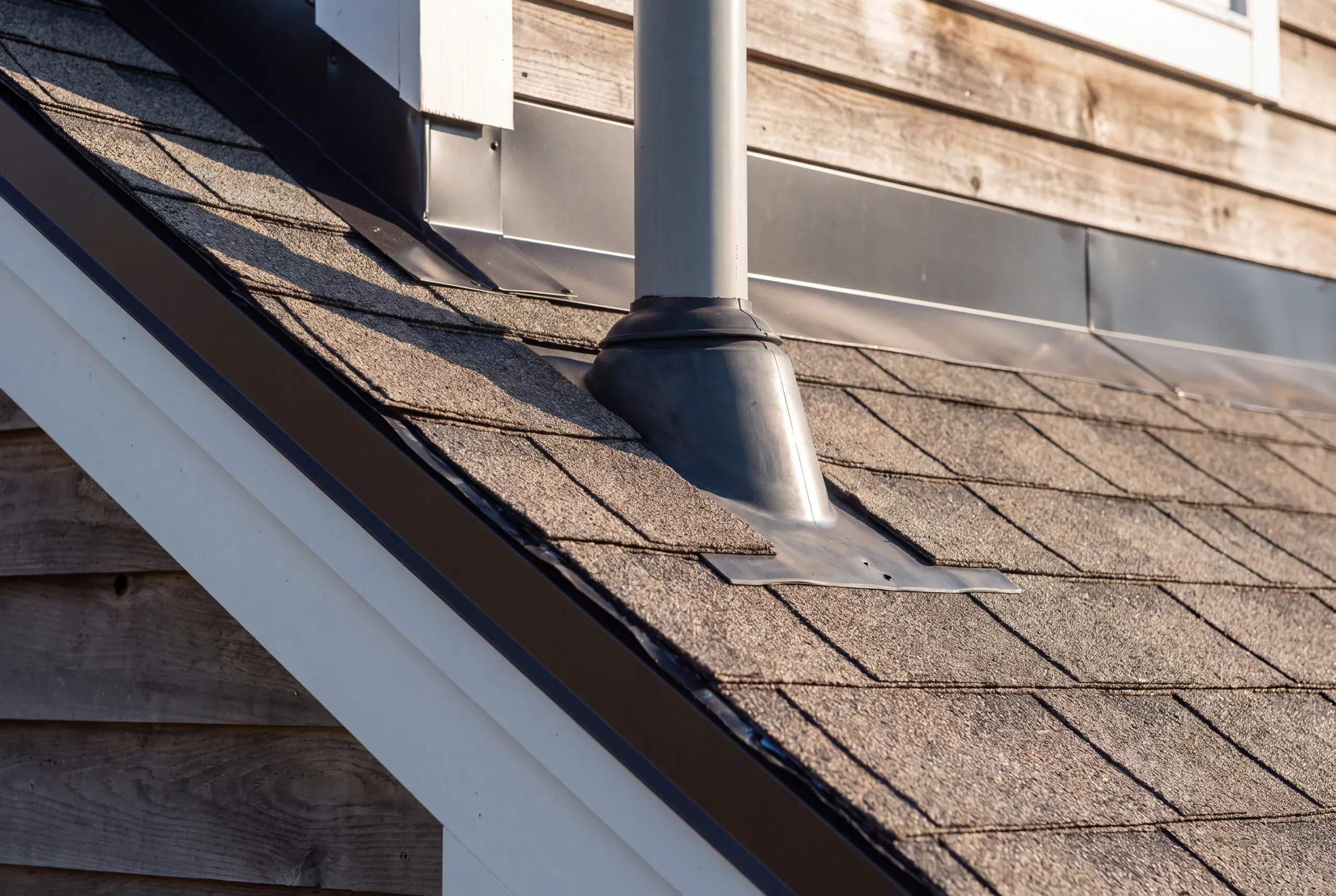Residential roofing contractor Pittston Maine: close view of a vent pipe boot on shingles beside metal step flashing and wood siding