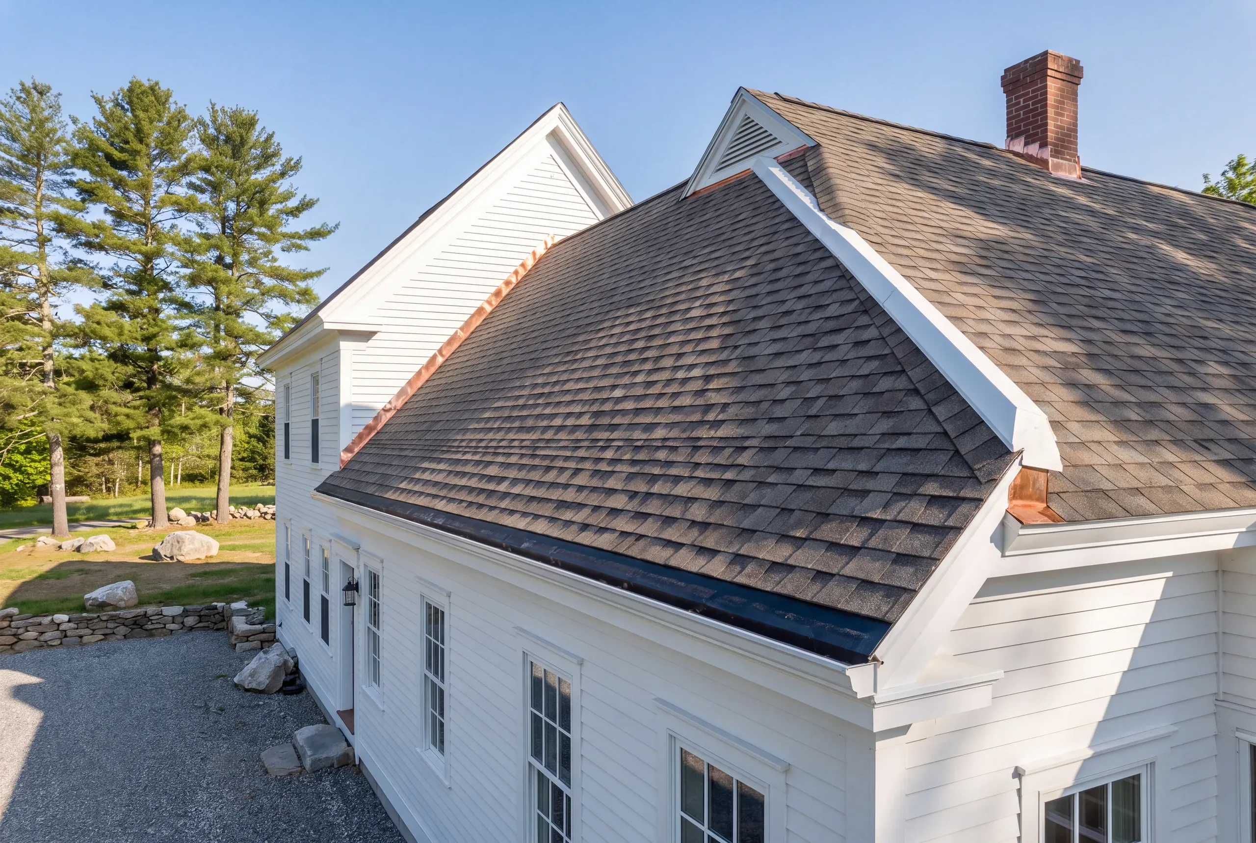 Residential roofing contractor Pittston Maine: white farmhouse with intersecting gables, brick chimney and stone wall by tall pines