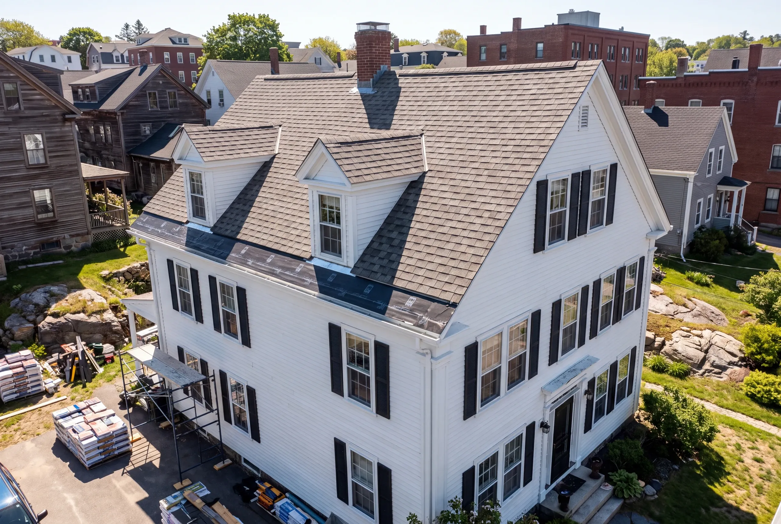 Roofing contractor in Rockland, ME: aerial view of a white colonial with gable dormers mid-reroof, scaffold and shingle pallets