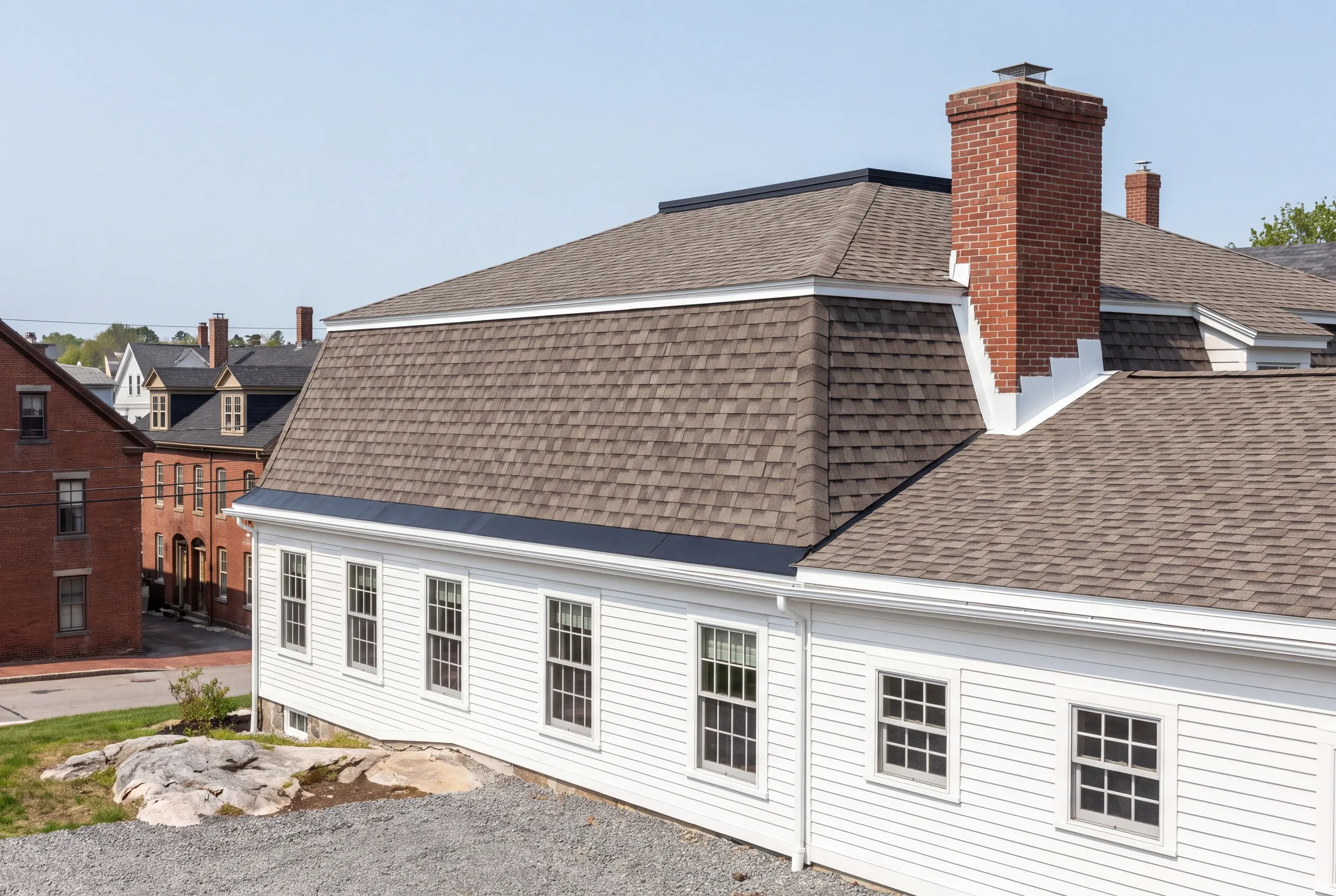 Residential roofing contractor Rockland Maine: long white house with a large brick chimney and two-level shingle roof on a ledge lot