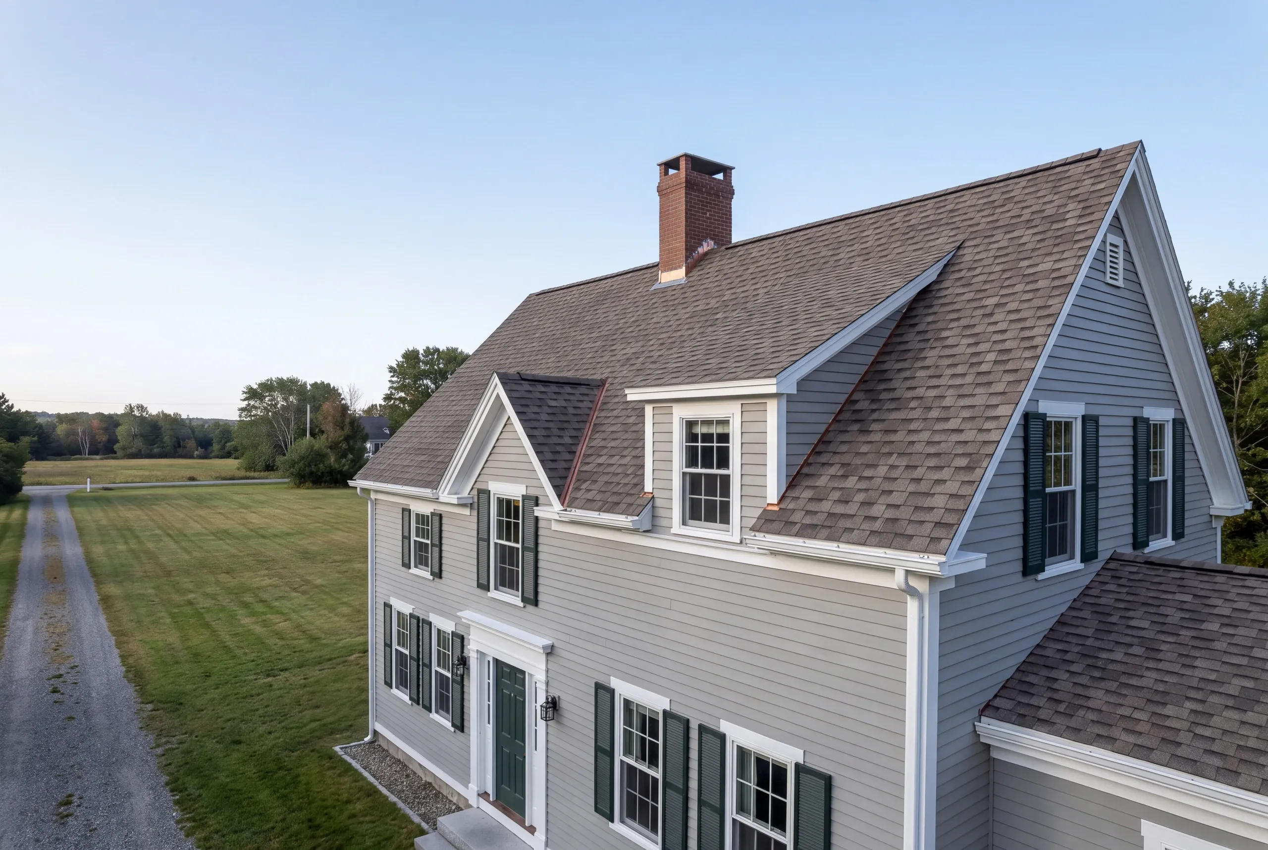 Roof replacement contractor Rockland Maine: gray-sided colonial with gable dormers, green shutters and a long gravel drive