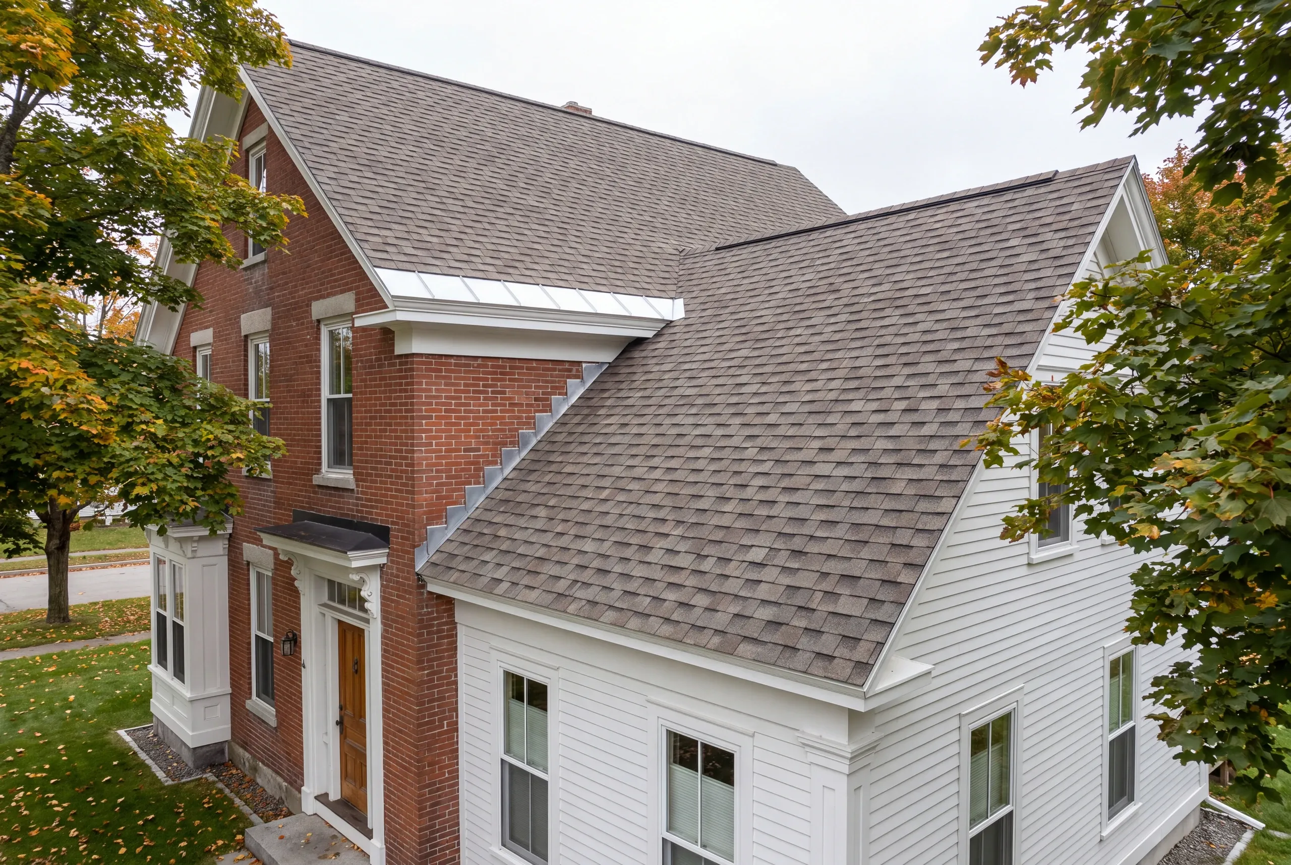 Roofing contractor in Waterville, ME: red brick house with a white clapboard ell, gray-brown architectural shingles and metal step flashing