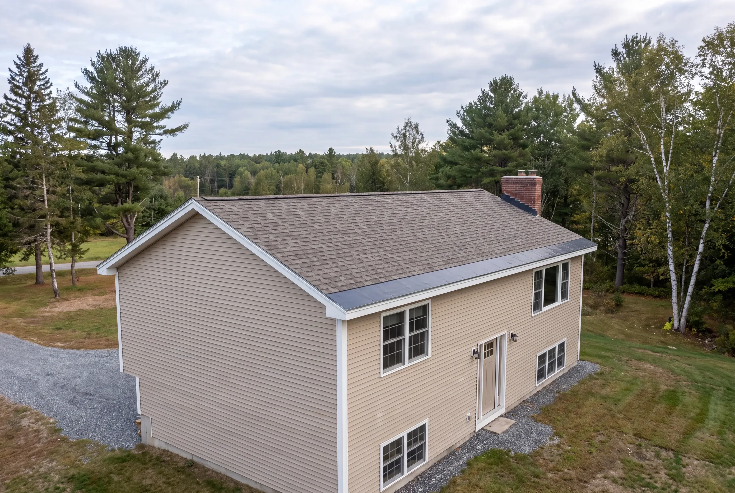 Roof replacement contractor Winslow Maine: split-entry house with tan lap siding, arch shingles, brick chimney and black metal eave band