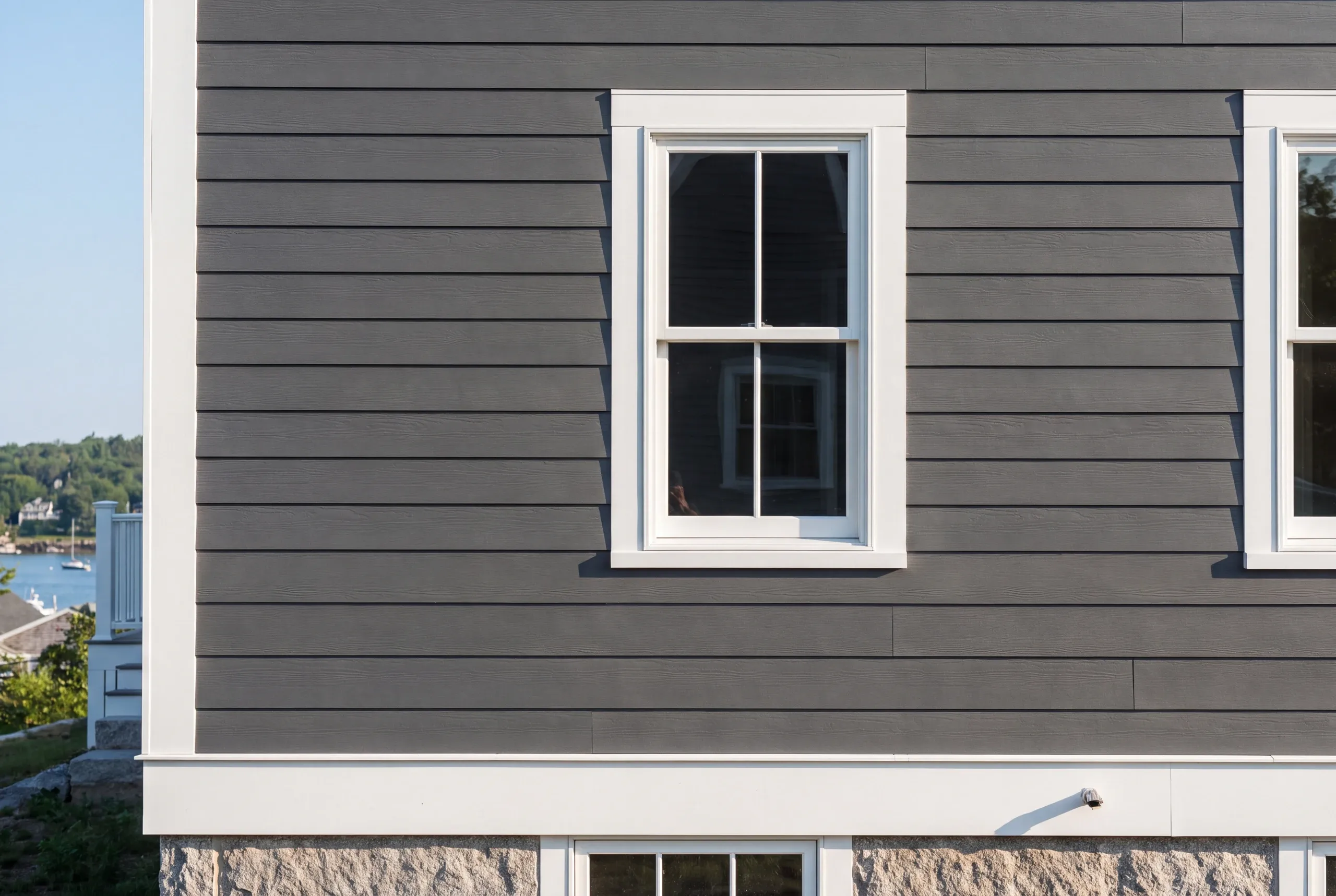 Siding installer Belfast Maine: charcoal wood-grain lap siding over a granite foundation with a harbor and sailboat beside