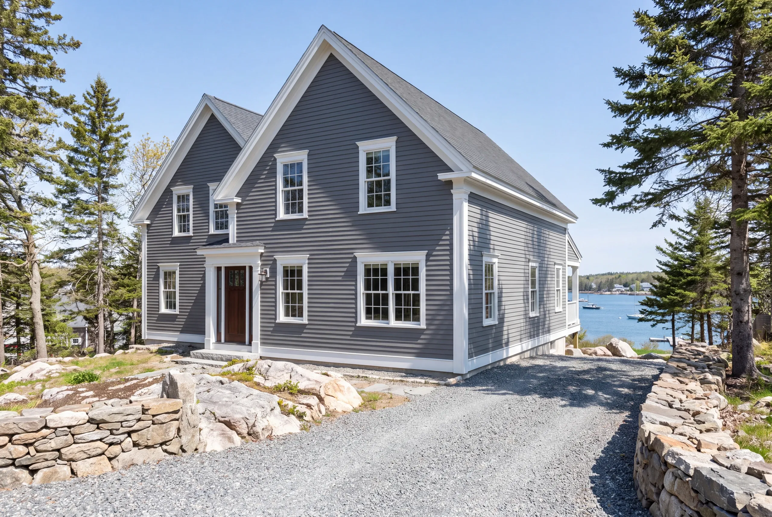 Exterior siding contractor Belfast Maine: gray lap-sided farmhouse with two front gables between stone walls above a harbor