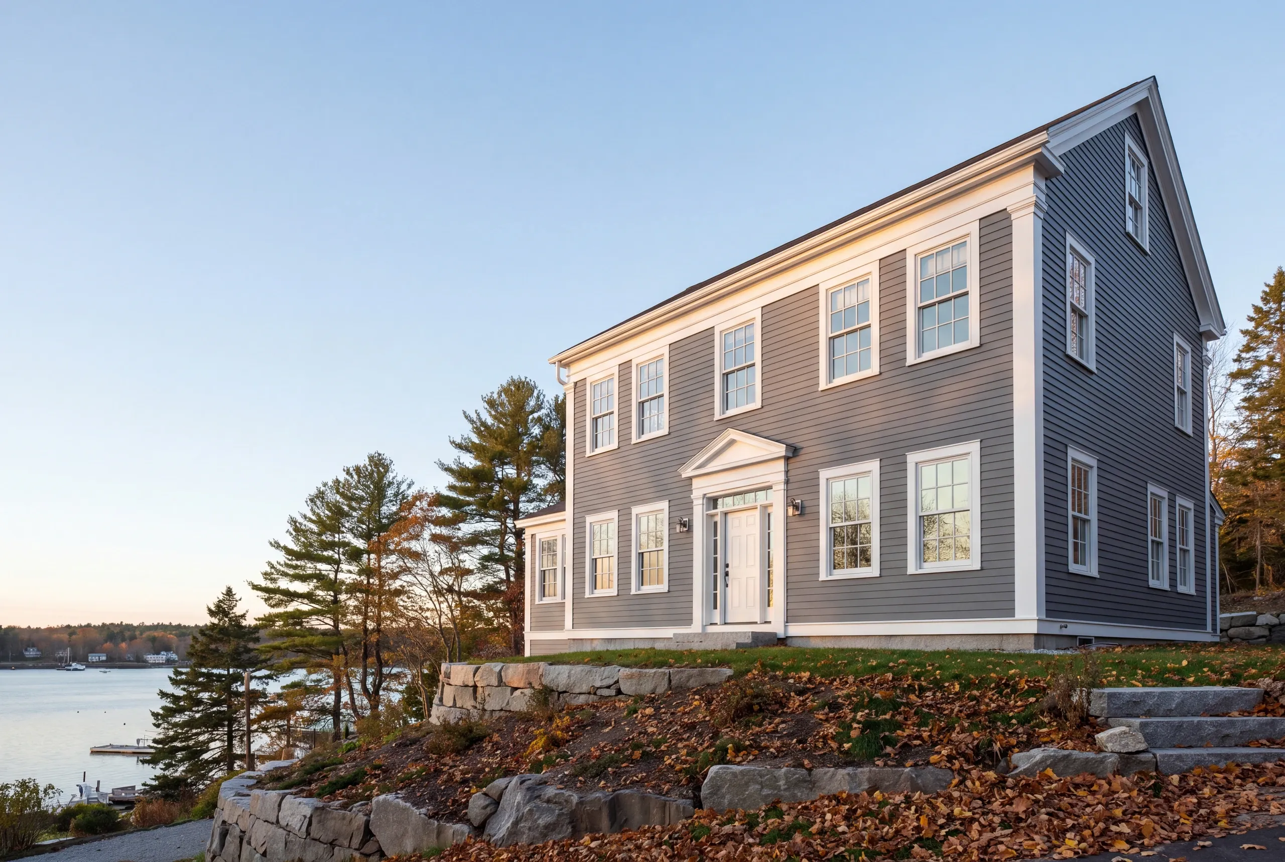 Exterior siding contractor Belfast Maine: warm gray lap-sided colonial with a pedimented entry above terraced granite walls and a harbor