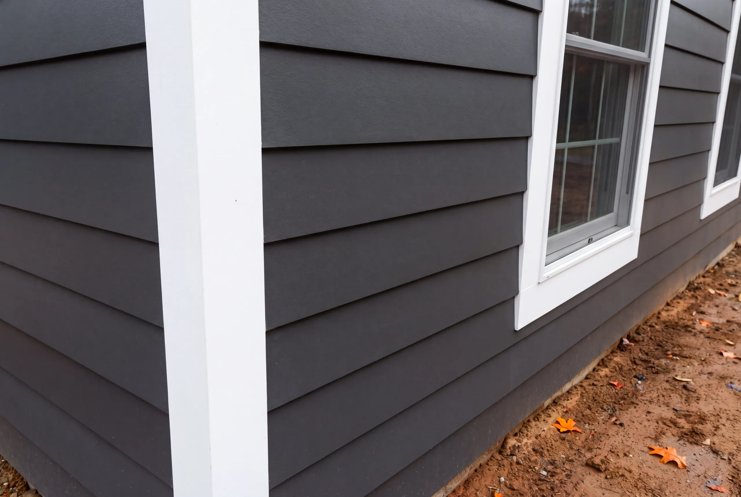 Siding installer Bucksport Maine: charcoal lap siding corner with a wide white corner board above raw dirt and fallen leaves