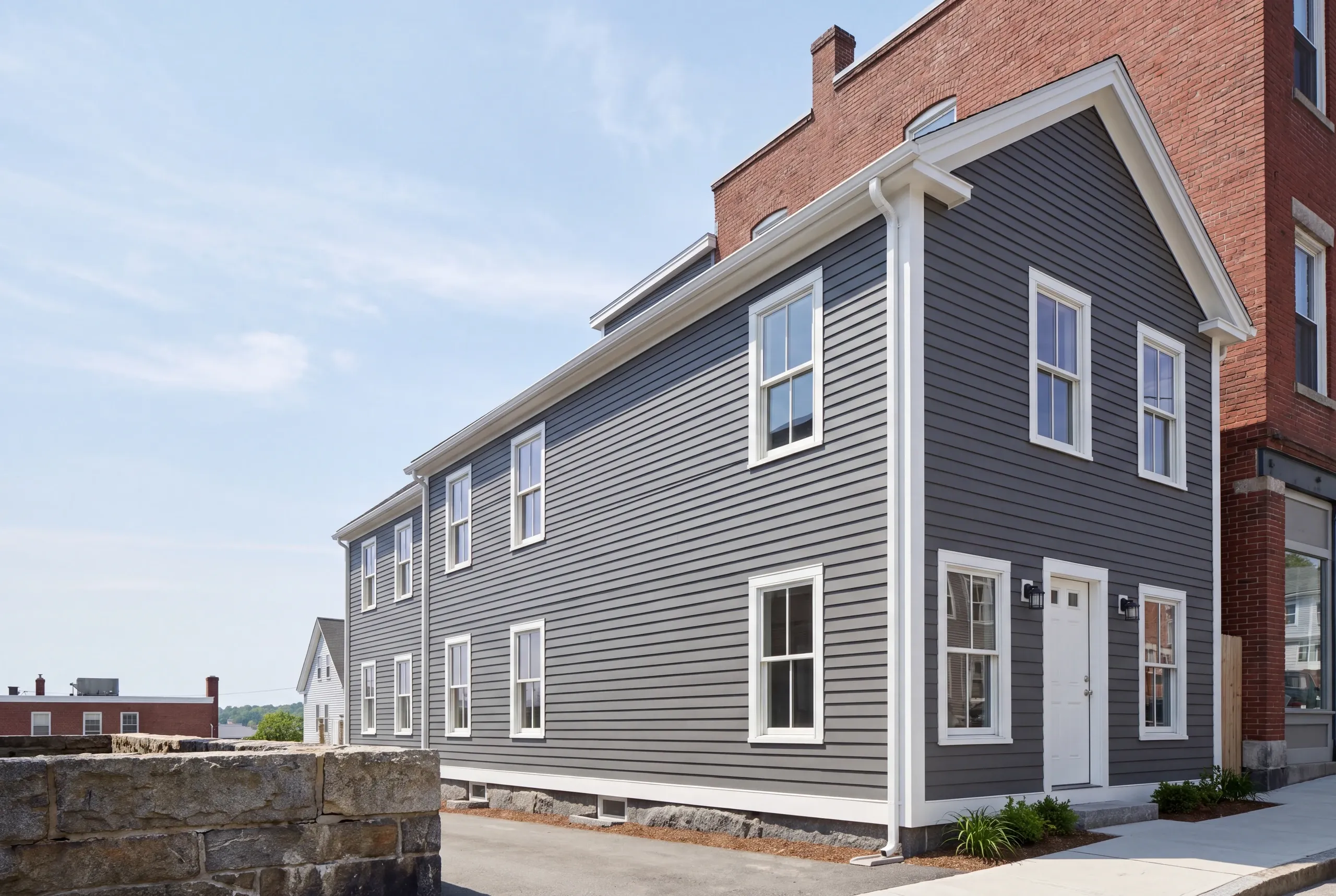 Exterior siding contractor Rockland Maine: two-story house in gray lap siding pressed against a tall brick building on a town sidewalk