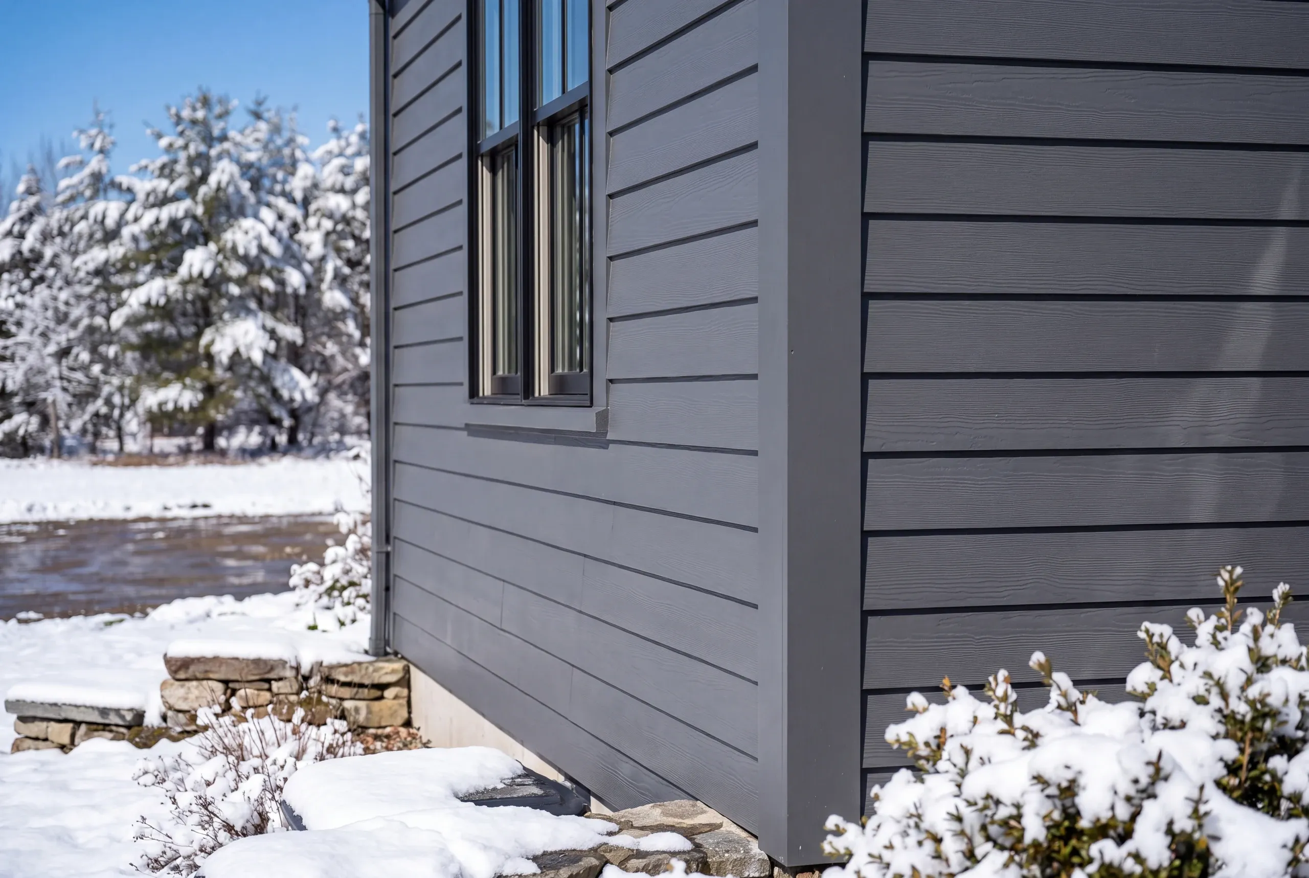 Siding installer Searsmont Maine: corner of a house in gray wood-grain lap siding with snow-covered shrubs and stone steps